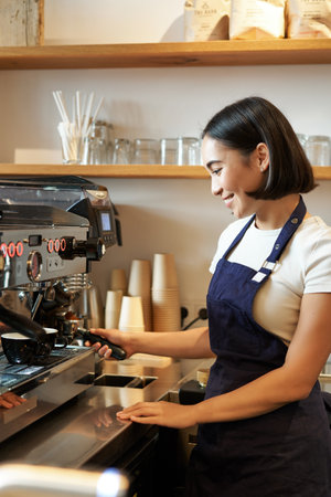 Happy beautiful asian girl, barista in apron works with coffee machine behind counter, standing in cafe and making cappuccinoの写真素材