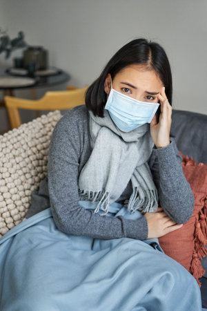 Covid-19 and health concept. Young asian woman in medical face mask, feels sick and unwell, catching flu, protecting others from influenza, sitting in living room covered with blanketの写真素材