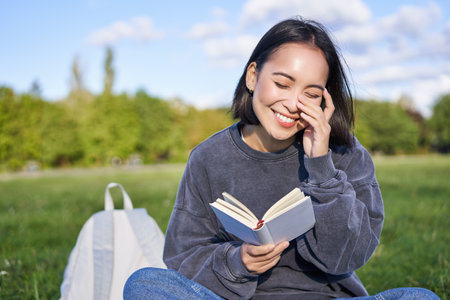 Beautiful woman sitting in park with book. Smiling asian girl reading and laughing, relaxing outdoorsの写真素材