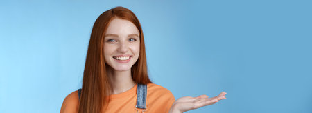 Proud good-looking confident redhead girl present awesome product hold object palm raise hand blank blue copy space smiling delighted recommend cool link, standing studio background helpfulの写真素材