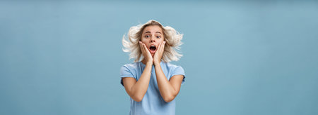 Indoor shot of shocked and impressed young stylish european female with blond hair holding hands on...