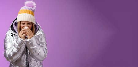 Girl runny nose sneeze tissue press napkin face got ill feeling unwell sick heading hospital standing purple background bending wearing silver stylish jacket winter hat, crying sobbingの写真素材