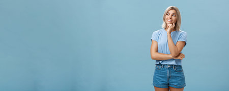 Dreamy thoughtful and creative artistic blonde woman in denim shorts and t-shirt smiling curiously...