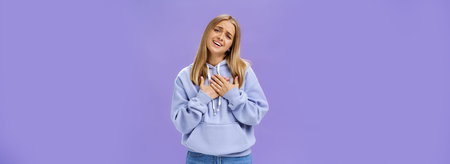 Sighing touched and pleased beautiful young girl with blond hair in hoodie holding palms on heart...