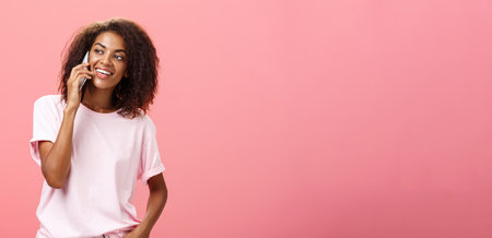 Girl calling friend to meet up. Portrait of charming friendly and outgoing african american young woman with afro hairstyle holding smartphone near ear while talking looking left curiouslyの写真素材