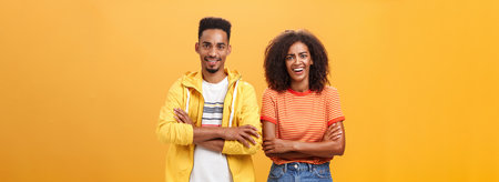 Portrait of surprised sarcastic african american woman with afro hairstyle standing with cute boyfriend crossing arms on chest laughing from scorn and fun over orange backgroundの写真素材