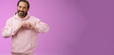 Family in heart. Portrait lovely handsome romantic bearded man in pink hoodie passionatly looking camera show love gesture smiling cute expressing romantic sympathy attitidue, purple backgroundの写真素材