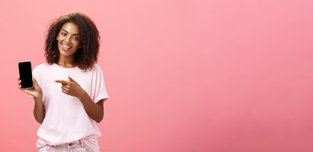 Suggest this model of phone. Happy charming african american curly-haired woman in stylish outfit holding smarpthone and pointing at device screen while recommending cool app over pink wallの写真素材