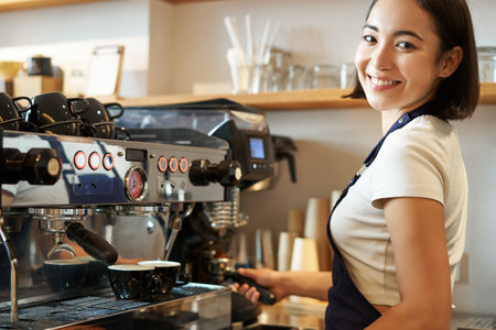Smiling asian woman working in cafe, using coffee machine for espresso, cappuccino orders, standing behind counter in blue apron, looking at cameraの写真素材