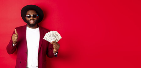 Handsome modern african american man in sunglasses and party clothes, showing thumb up with dollars, earn money and looking satisfied, red backgroundの写真素材