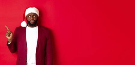 Christmas, party and holidays concept. Skeptical and gloomy african american man pointing finger left, looking disappointed, standing in santa hat against red backgroundの写真素材