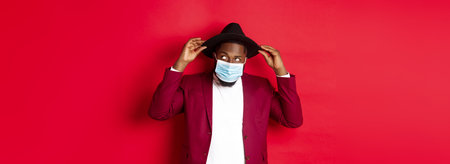 quarantine and holidays concept. Handsome and stylish african american man in face mask, put hat on...