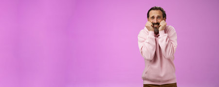 Scared insecure silly adult bearded man grey hair in pink hoodie press palms mouth bite fingers...