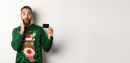 Christmas holidays and shopping concept. Man showing credit card and looking surprised, standing over white backgroundの写真素材