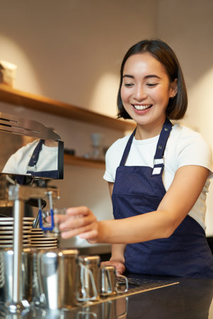 Portrait of cute barista girl working behind counter, making coffee, steaming milk for cappuccino, wearing cafe uniform blue apronの写真素材