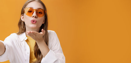 Stylish popular girl sending air kiss at camera taking selfie or recording vlog working as freelance...