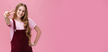Charismatic young teenage girl in corduroy overalls posing joyfully and energetic against pink background showing peace sign at camera holding hand on waist in confident pose and smiling cheerfullyの写真素材