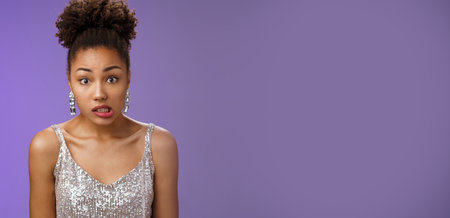 Awkward cute african-american woman in silver glamour dress clench teeth look worried complicated say oops make mistake accidently spill drink standing unsure insecure blue backgroundの写真素材