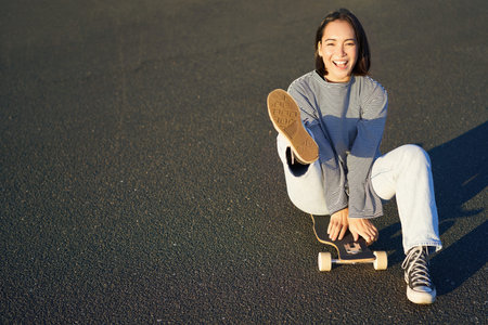 Portrait of beautiful asian girl skating, sitting on her skateboard and smiling. Cute teenager with...