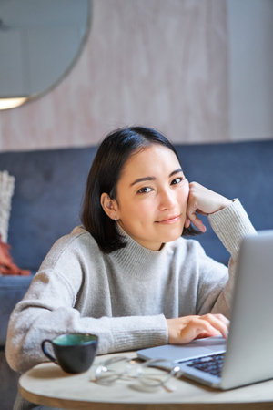 Vertical shot of young asian woman looking tired, smiling with exhausted expression, working from...