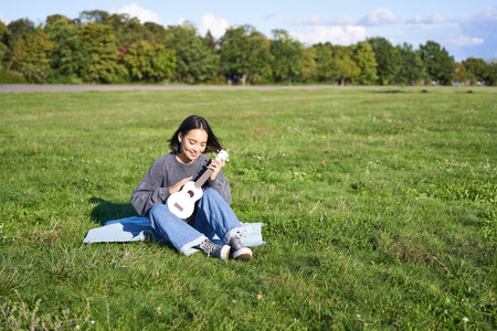 Portrait of asian girl sitting alone in park, playing ukulele and singing, enjoying leisure time