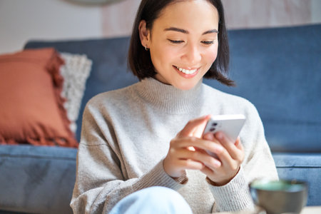 Technology and people. Young stylish asian woman sits at home with her smartphone, texting message, using application on mobile phoneの写真素材