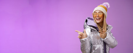 Hey how you doin. Cheeky stylish bond girl having fun greeting friend pointing finger pistols left smiling sassy say hello what up wearing cool silver jacket hat sunglasses, purple backgroundの写真素材