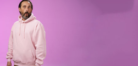 Impressed speechless shocked adult bearded man grey hair in cool pink hoodie folding lips wow popping eyes camera surprised astonished intrigued, standing curious amazed purple backgroundの写真素材