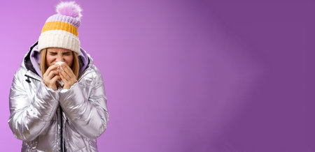 Girl runny nose sneeze tissue press napkin face got ill feeling unwell sick heading hospital standing purple background bending wearing silver stylish jacket winter hat, crying sobbingの写真素材