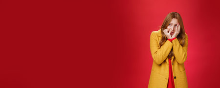 Portrait of shocked and scared insecure woman in yellow coat covering face in horror and fear biting...