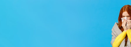 Seasonal flu. Cute redhead female student cought cold, sneezing in napkin, wear sweater and scarf on shoulders, feeling unwell sick, suffering high temprature, standing blue backgroundの写真素材