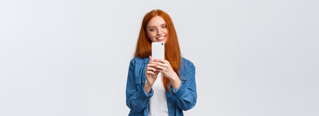 Waist-up portrait cheerful modern cute redhead woman taking photo of friend, smiling and tilt head lovely, looking mobile display, shooting beautiful scenery to post online in blog, white backgroundの写真素材