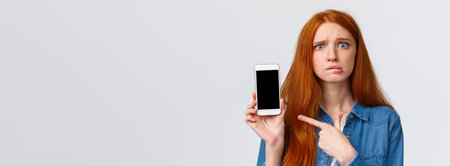 Worried, insecure and alarmed cute redhead girl asking for advice having problems messaging with stranger on dating app, dont know what answer standing nervous pointing finger at smartphone displayの写真素材