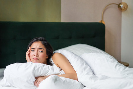 Sad and distressed young korean woman lying in her bed with pillow and warm duvet, feels uneasy, frowning and grimacingの写真素材