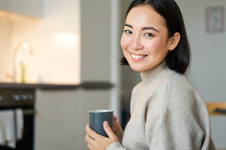 Portrait of smiling asian girl with cup of coffee, drinking hot tea and getting cozy at home. Warming up with beverageの写真素材