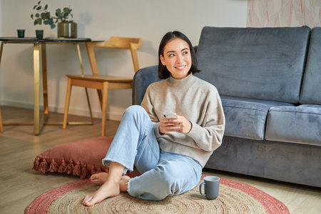 Image of stylish young woman in modern house, using mobile phone, sitting on floor and holding smartphone, drinking from cupの写真素材