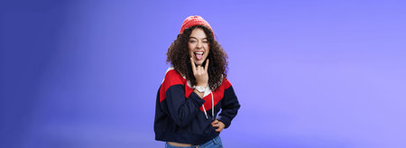 Stylish and cool energized swag girl with curly hairstyle in warm beanie showing rock-n-roll gesture...