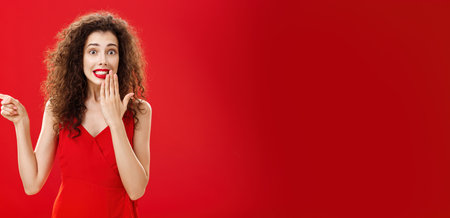 Troubled cute and tender silly woman with curly hairstyle in red stylish evening dress saying oops smiling with guilty look covering mouth with palm while pointing left over studio backgroundの写真素材