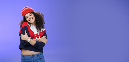 Sweet tender and sensual curly-haired woman in red stylish beanie hugging herself with closed eyes...
