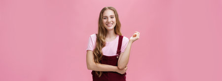 Cute girl in trendy overalls with long wavy hairstyle and light makeup standing in casual pose and smiling cheerfully at camera listening taking part in conversation with pleasant positive attitudeの写真素材