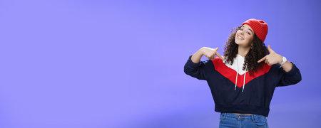 Proud and self-satisfied happy young energized stylish female model in warm beanie brag about own achievements as pointing at herself raising head and smiling produly posing delighted over blue wallの写真素材