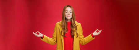 Girl keep calm releasing stress with meditation, posing in yellow coat, close eyes and looking relieved as extending hands sideways with mudra gesture, doing yoga against red background in lotus poseの写真素材