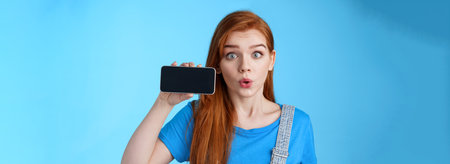 Amazed fascinated excited 20s redhead woman show smartphone screen horizontally, fold lips whistling amused wondered, check out cool phone app, stand blue background curiousの写真素材