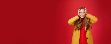 Portrait of redhead woman in panic holding hands pressed to head clenching teeth and frowning puzzled and troubled, standing anxious and concerned freaking out over red backgroundの写真素材