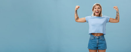 Strong women stand up and fight for rights. Portrait of happy entertained and cool young female athletic blonde with tattoos winking and smiling showing muscles and biceps over blue wallの写真素材