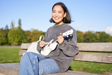 Happy cute girl sits alone on bench in park, plays ukulele guitar and enjoys sunny day outdoorsの写真素材