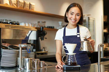 Smiling girl barista, asian bartender pouring water from kettle, brewing filter coffee in cafe behind counter, wearing blue apronの写真素材