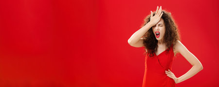 Woman being pissed with dumb people. Irritated forgetful adult caucasian female with curly hair in red elegant dress punching forehead with palm closing eyes and swearing recalling important thingの写真素材