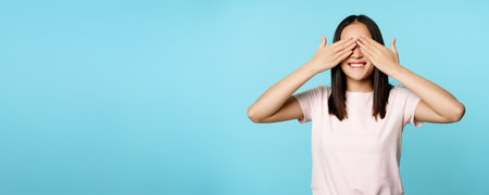 Happy smiling asian girl covers ears and waits for surprise, standing in t-shirt over blue backgroundの写真素材