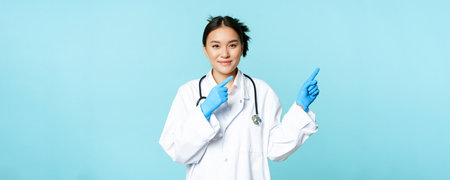 Smiling asian healthcare worker, wearing sterile gloves and uniform, pointing right, demonstrating chart, hospital advertisement, blue backgroundの写真素材